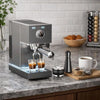 A stainless steel espresso tamper and dosing kit sits on a countertop next to coffee beans and a white mug.