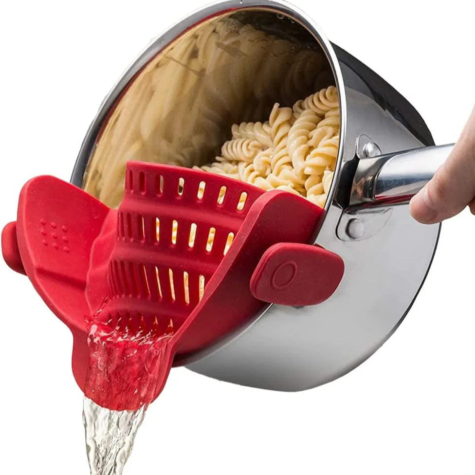 A red silicone clip-on strainer is pouring water over dry pasta inside a metal colander.