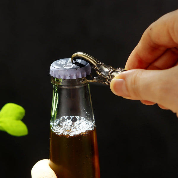 A hand is using an Artisan Scarab Beetle bottle opener to open a glass bottle containing dark liquid.