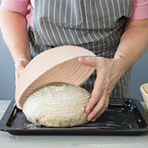 A person is holding a natural rattan bread proofing basket over a loaf of bread on a baking tray.