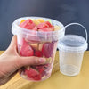 A hand holds a clear plastic storage bucket filled with sliced strawberries and other fruit, next to an empty clear plastic c