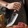 A hand holds a forged chef knife with a wooden handle, resting on a wooden board next to raw meat.