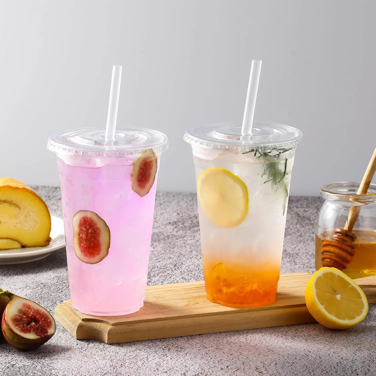 Two clear plastic cups filled with colorful iced beverages, garnished with citrus slices and herbs, resting on a wooden board