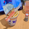 A plastic cup filled with noodles and a pink squid-shaped fork, set against a blurred background of a laptop.