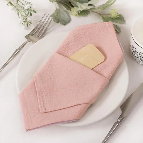 Ein Set gefalteter rosa wiederverwendbarer Baumwollservietten auf einem weißen Teller mit Besteck und Grünzeug.