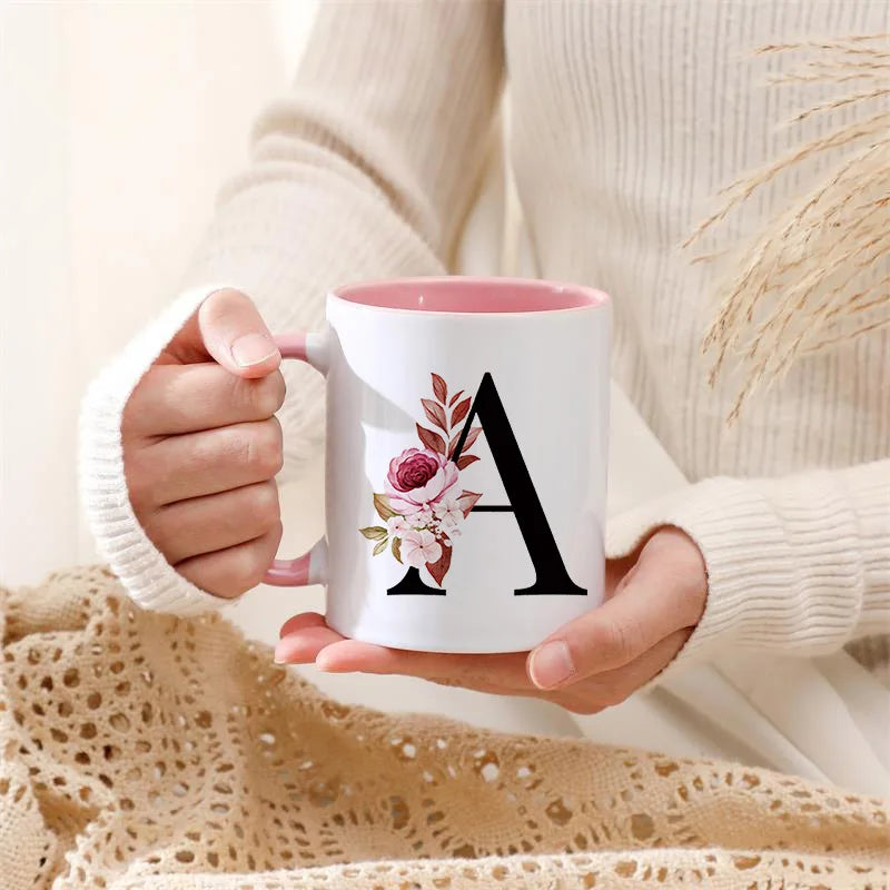 A white coffee mug with a large black letter A and pink floral accents is held by a person wearing a cream sweater.