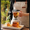 A hand pours brewed coffee from a glass dripper into a mug, set on a wooden tray.