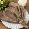 A stack of six brown cloth napkins tied with twine is displayed on a white plate next to green foliage and white candles.