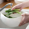 A hand is using a kitchen strainer to pour water over fresh green leaves in a white bowl.