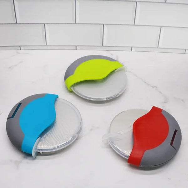 Ein Set aus drei Plastik-Pizza-Scheren in Grau, Limettengrün und Rot auf einer weißen Marmor-Arbeitsplatte.
