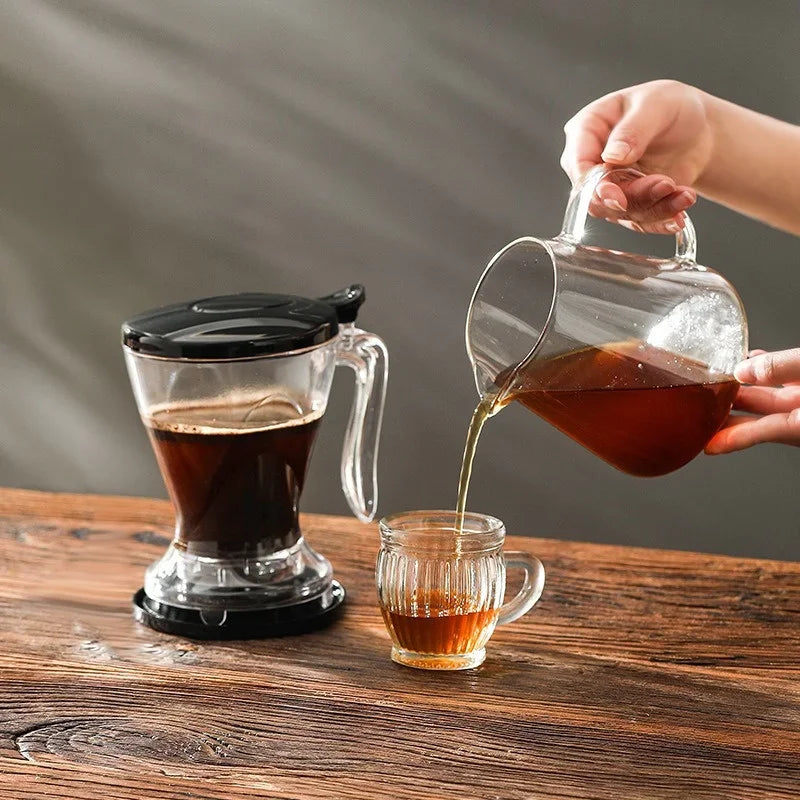 A hand pours brewed coffee from a glass dripper into a small cup on a wooden table.