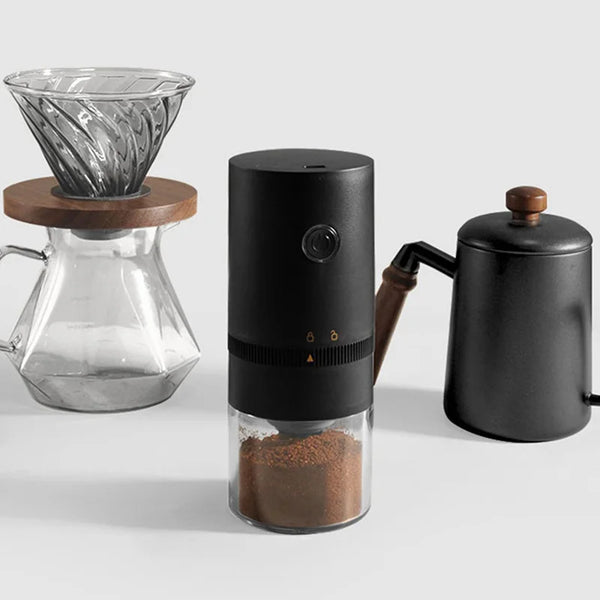 A black portable USB-C burr coffee grinder is shown next to a glass grinder and a dark coffee pot.