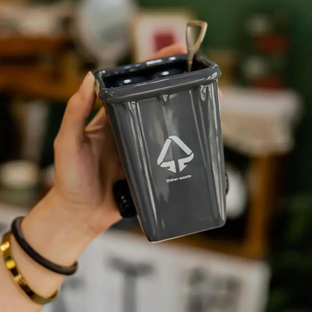 A hand holds a dark gray novelty trash can with a gold spoon inside, set against a blurred background.