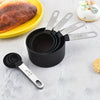 A black stainless steel measuring cup and spoon set with various sizes is displayed on a white surface next to a baked good.