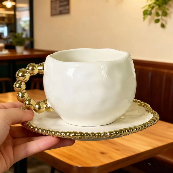 Weiße Keramiktasse mit goldperlenhenkel auf passender goldrandiger Untertasse.