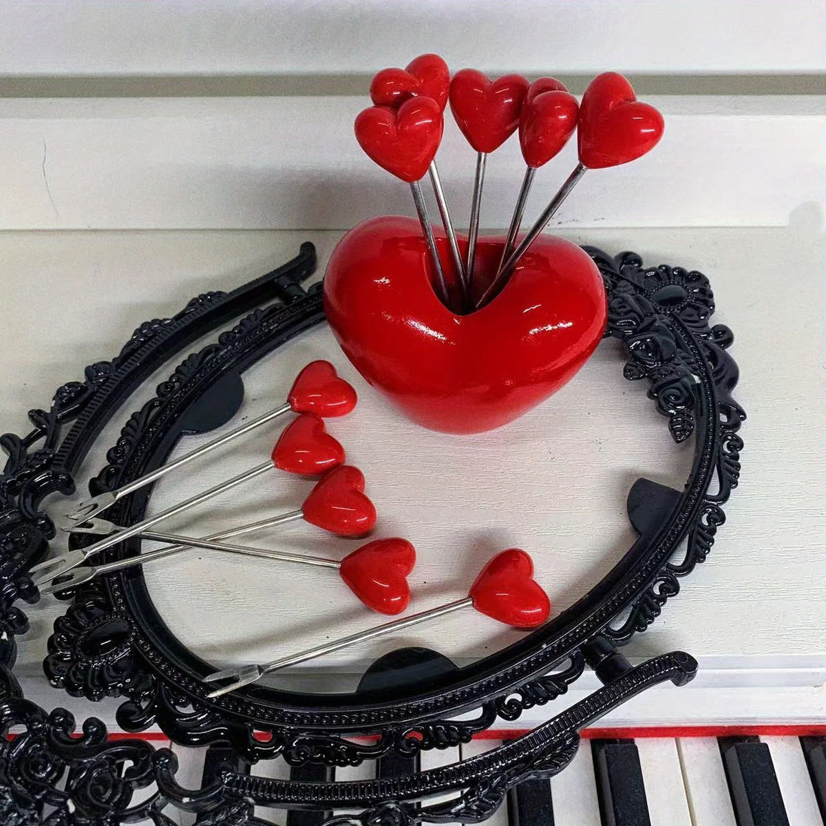 A set of red heart-shaped stainless steel fruit forks arranged around a black decorative wreath on a piano keyboard.