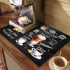 A black coffee drying mat is placed on a wooden table next to a silver coffee machine and a cup of coffee.
