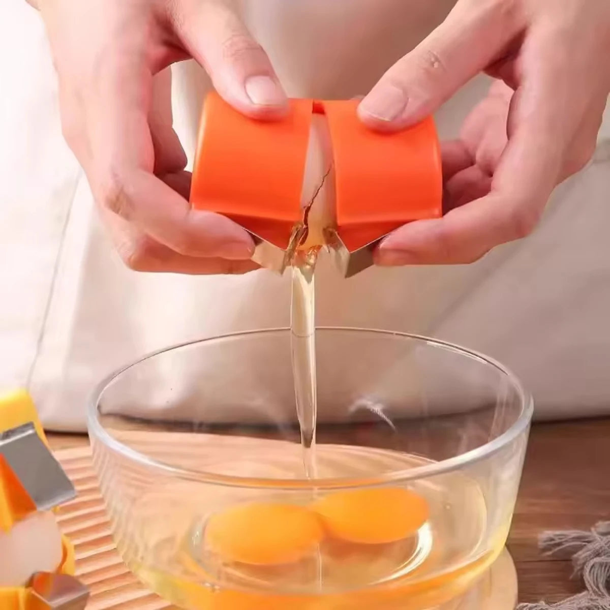 Hands are using a portable egg shell opener and separator tool to crack an egg in a glass bowl.