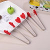 Red silicone food tongs with heart shapes are displayed on a white plate next to a citrus fruit and a metal tray.