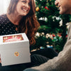 A woman smiles while holding a white box with a thank you note, set against a blurred Christmas tree background.