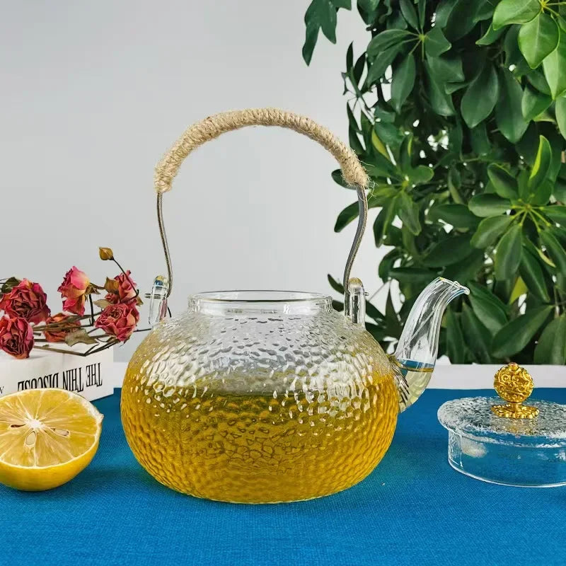 Hand-hammered glass teapot with a hemp rope handle filled with yellow tea on a blue cloth.