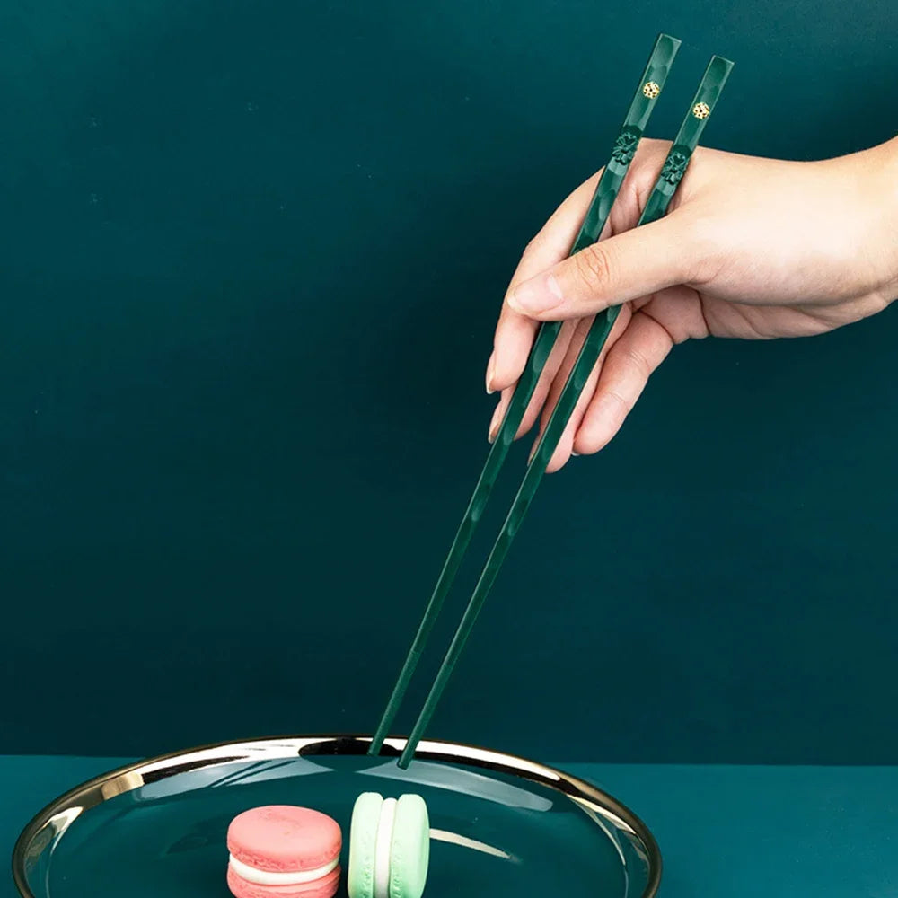 A hand holds a pair of green reusable chopsticks over a glass bowl containing pink and green food items.