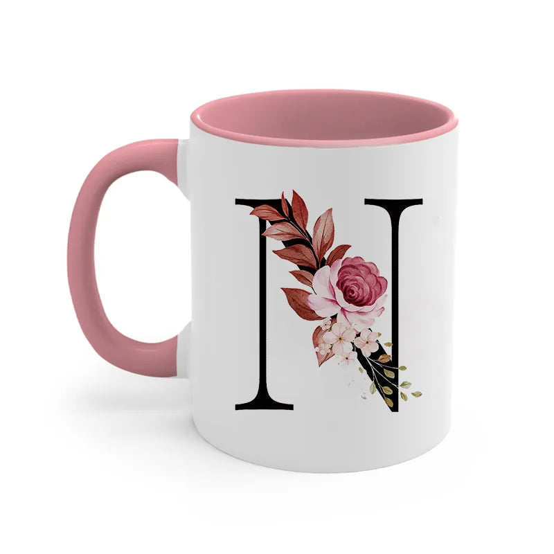 A pink ceramic coffee mug with a white background featuring a large black initial 'I' decorated with floral and leaf elements