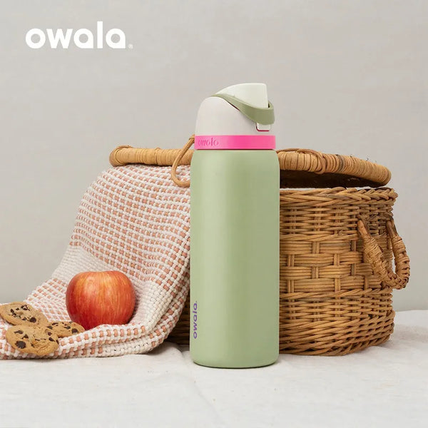Helles grünes Owala isoliertes Edelstahl-Travel-Glas mit rosa Deckel neben einem geflochtenen Korb und einem Apfel.