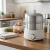 A silver electric steamer for eggs sits on a wooden countertop next to raw eggs and a cutting board.