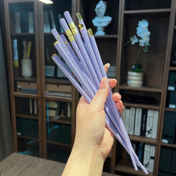 A hand holds a set of purple reusable anti-slip dining chopsticks against a wooden bookshelf background.