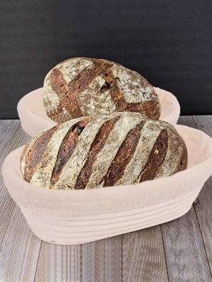 A loaf of dark bread with striped crusts is placed in a natural rattan bread proofing basket.