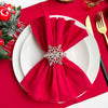A set of six burgundy cloth napkins tied in a bow on a white plate, set against a red tablecloth with festive greenery.