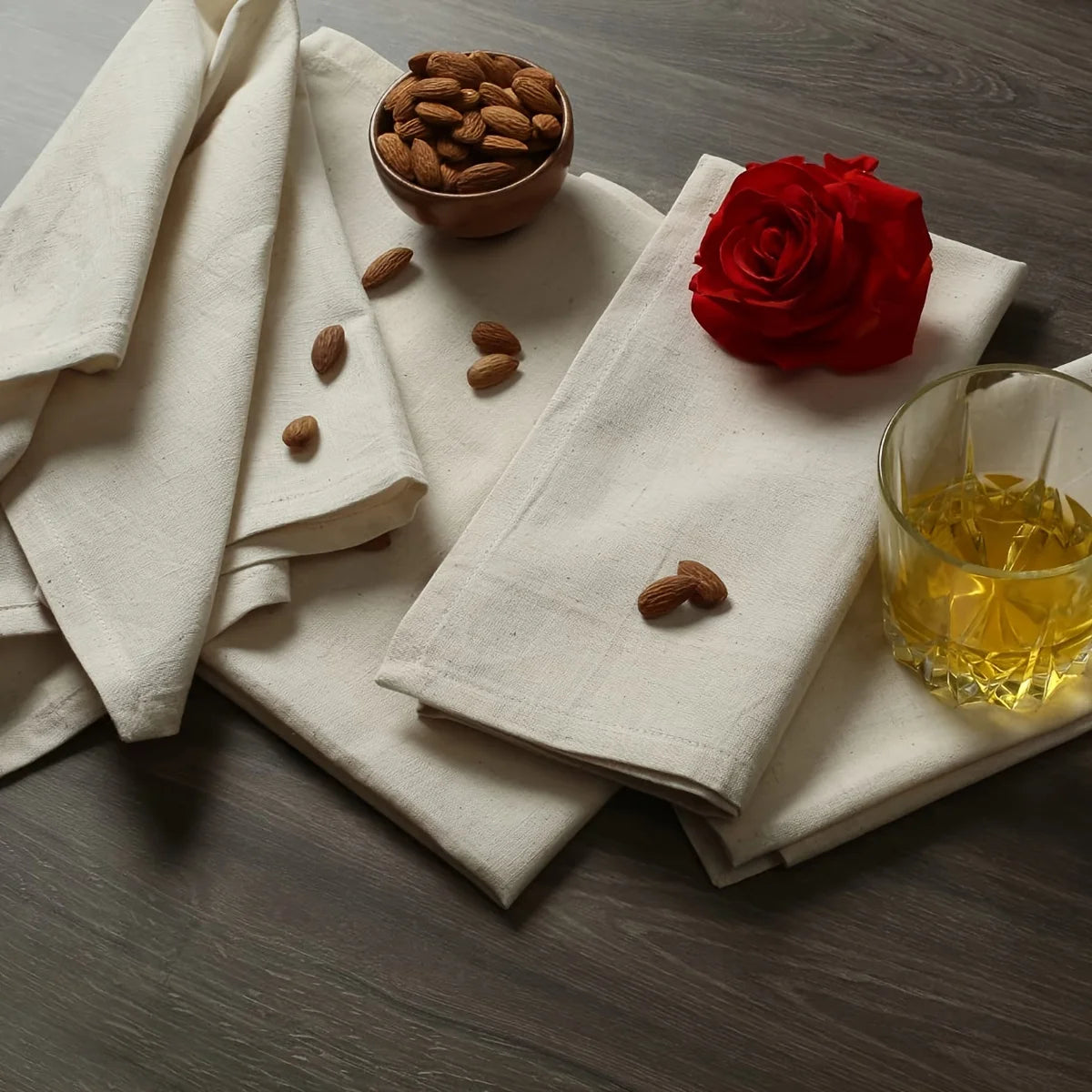 A set of white decorative table napkins is displayed on a dark wooden surface with a red rose and some almonds.