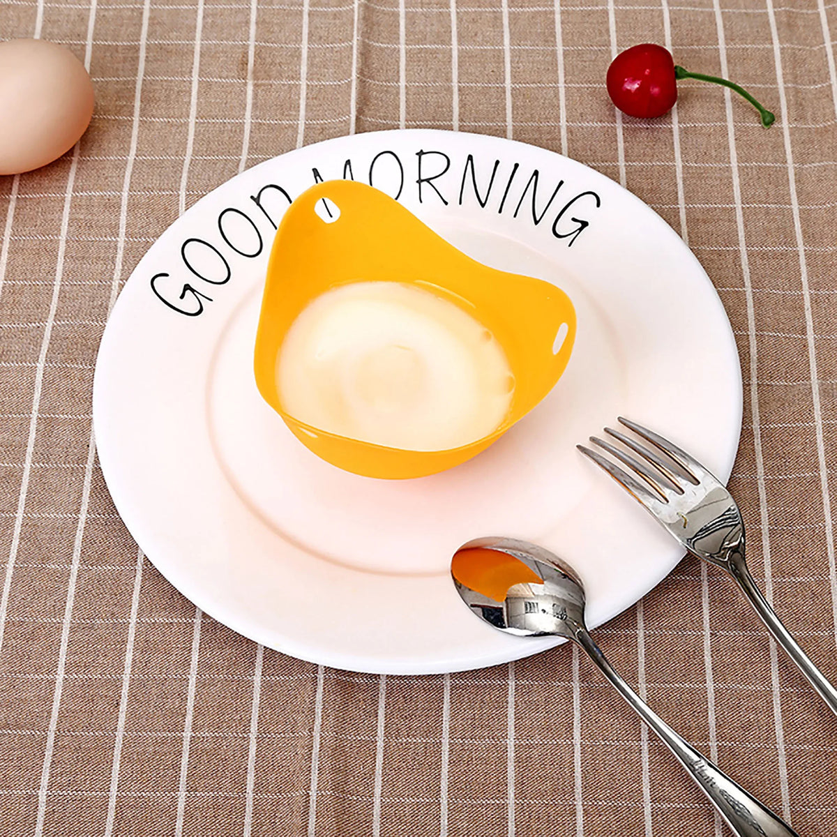 A white plate with a yellow silicone egg poacher mold, silverware, and a cherry on a beige checkered cloth.