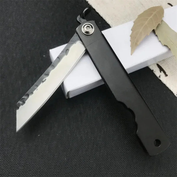 Schwarzes faltbares Taschenmesser mit silberner Klinge auf dunkler Oberfläche neben weißer Box und trockenem Blatt.
