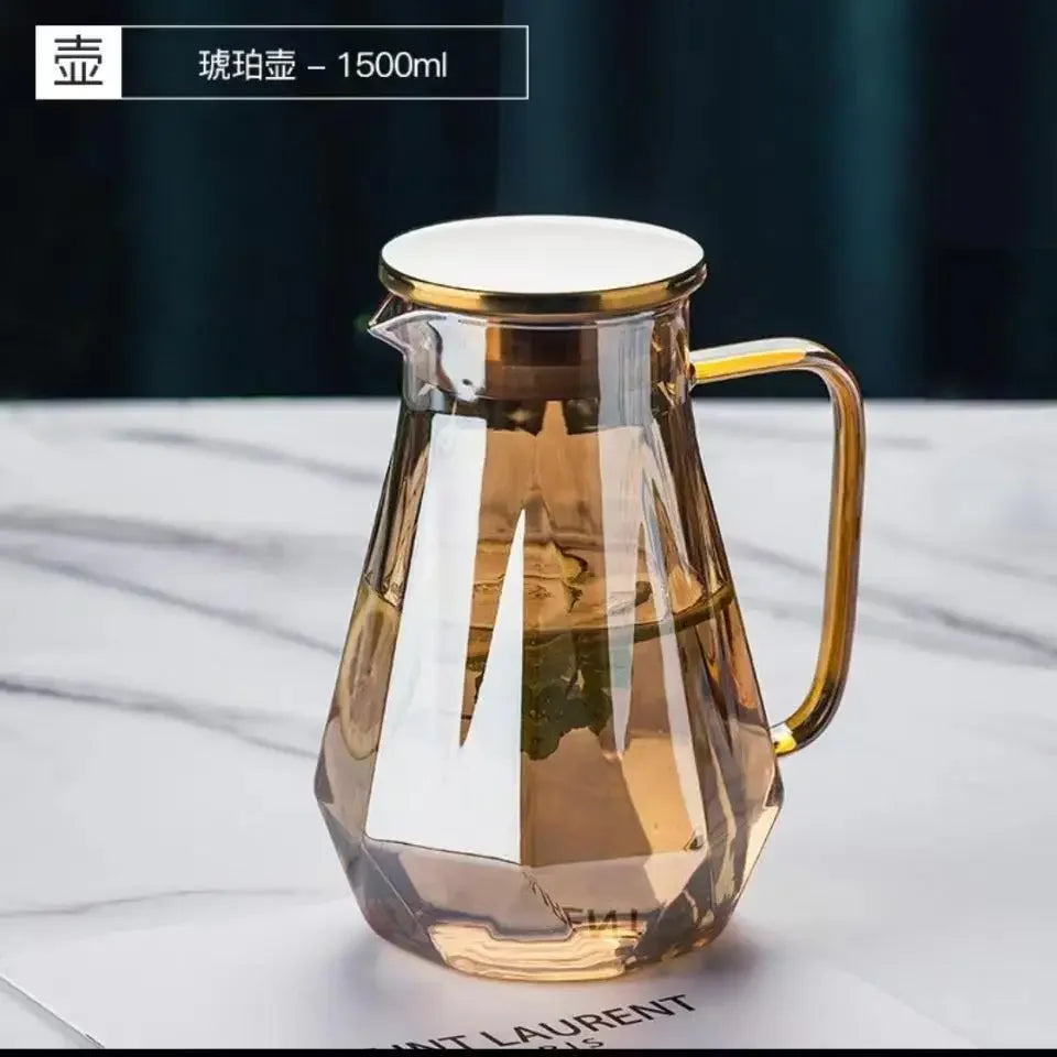 A clear glass pitcher with a gold rim and handle, filled with amber liquid, set against a white background.