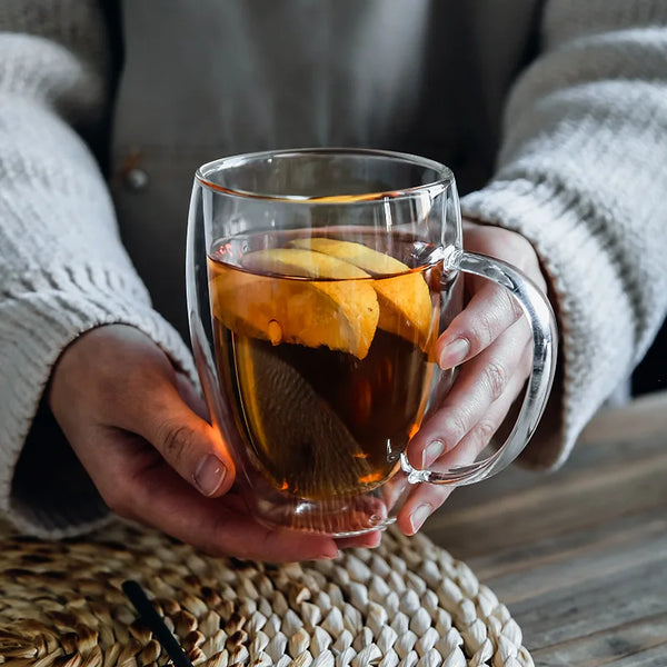 Eine Person hält eine Glas-Kaffeetasse mit dunkler Flüssigkeit und Zitronenscheiben vor einem unscharfen Hintergrund.