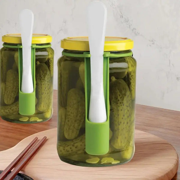 Ein Glas-Cornichonglas mit grünem Deckel und weißem Gabelgriff auf einem Holzbrett.