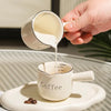 A hand pours milk from a white ceramic cup into another white ceramic coffee cup on a light tray.