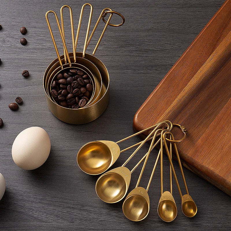 A set of gold-toned stainless steel measuring cups and spoons arranged on a dark surface with coffee beans and an egg.