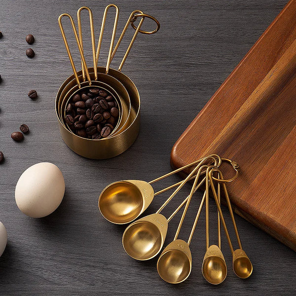Set goldfarbener Edelstahlmessbecher und -löffel mit Kaffeebohnen und einem Ei auf dunkler Oberfläche