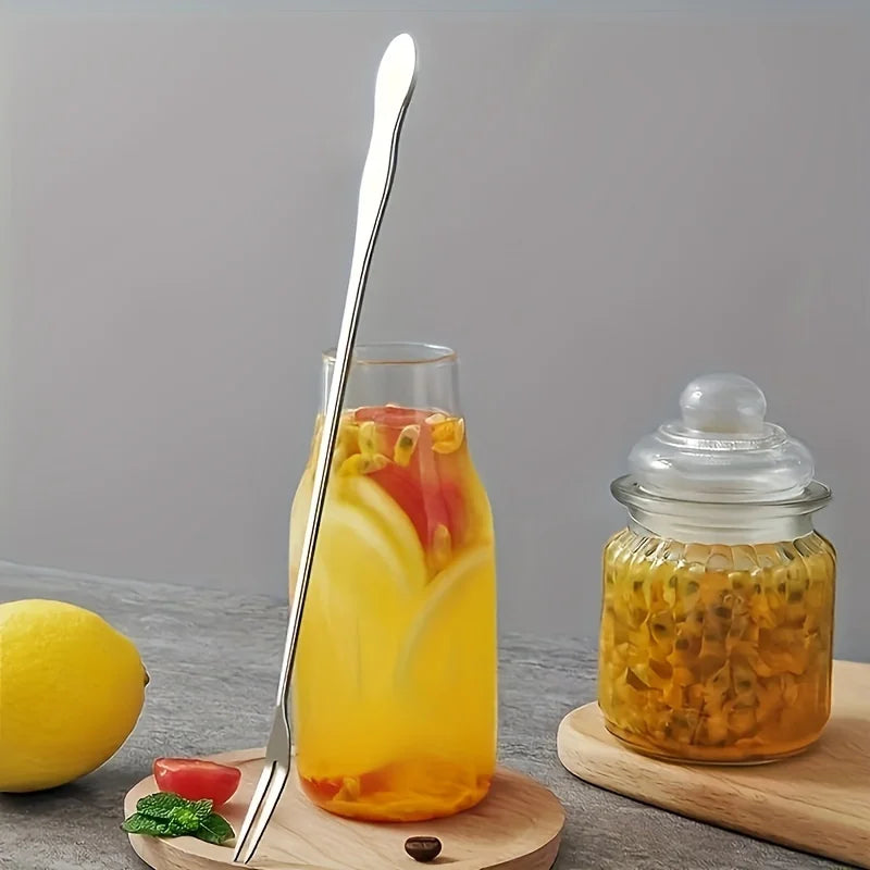 A stainless steel fork is placed next to a glass of yellow juice and a jar of pickled fruit on a wooden surface.