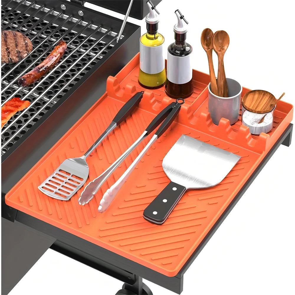 An orange silicone grill mat with various metal cooking utensils and a drip pad set on a black grill.