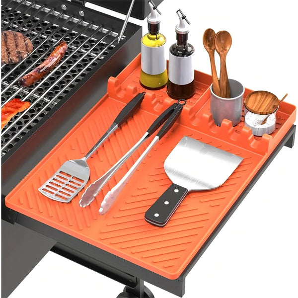 An orange silicone grill mat with various metal cooking utensils and a drip pad set on a black grill.