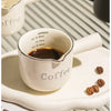 A white ceramic espresso coffee cup with a handle and a measuring scale is shown on a light plate.