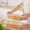 A person is preparing food using a clear plastic container filled with noodles on a wooden table.
