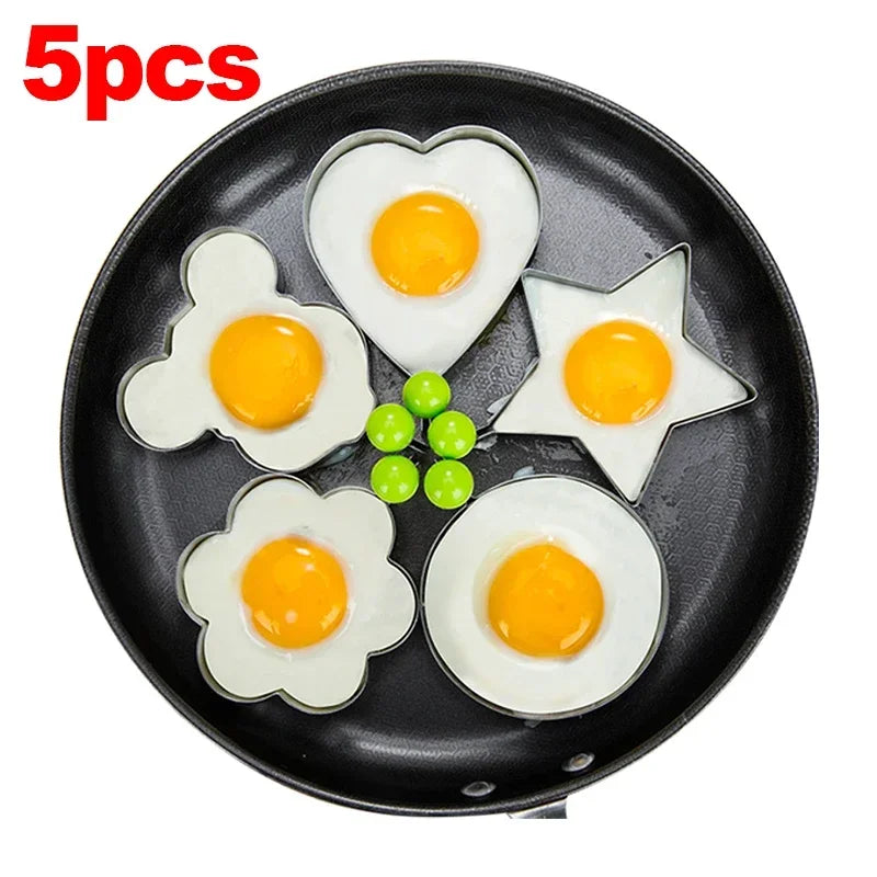 Five stainless steel fried egg molds are arranged on a black frying pan.