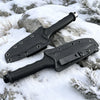 A black fixed blade utility knife with a nylon sheath rests on a snowy rock outdoors.