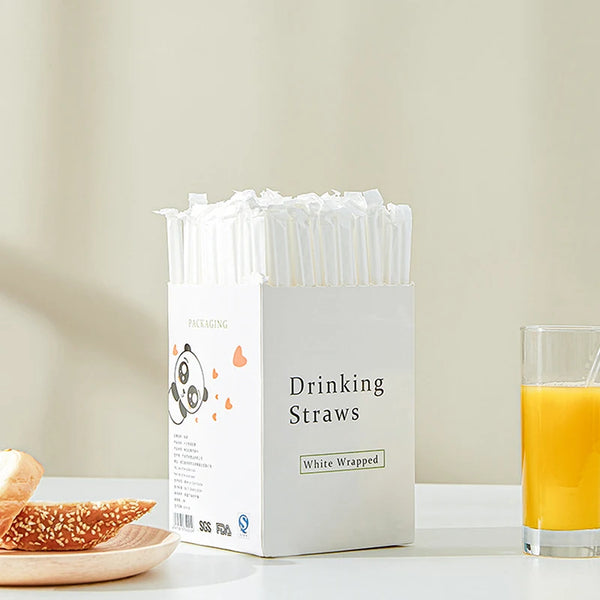 A white box of individually wrapped clear plastic drinking straws is displayed next to a glass of orange juice.