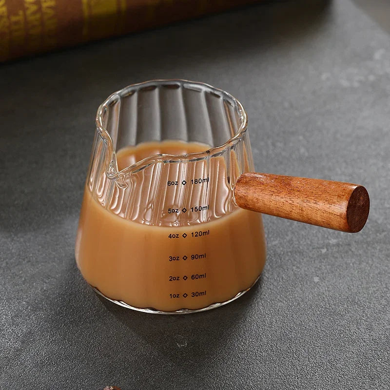 Taza medidora de espresso de vidrio preciso con mango de madera mostrando varias medidas líquidas.
